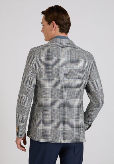 Hombre vistiendo un blazer a cuadros gris y azul y pantalones azul marino, de espaldas sobre un fondo liso gris claro.