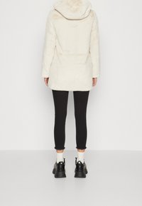 Abrigo de piel sintética color crema con capucha, que presenta mangas largas y un corte por encima de la rodilla, combinado con pantalones slim fit negros y zapatos negros gruesos.