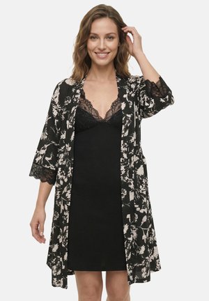 Donna che indossa un vestito nero con pizzo e una vestaglia a fantasia floreale bianca e nera, sorridente con la mano che tocca i capelli.