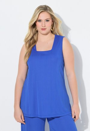 Blondynka w złotych kolczykach, ubrana w niebieską bluzkę bez rękawów i dopasowane niebieskie spodnie, stoi na białym tle.