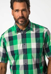 Camisa de cuadros de manga corta en verde, blanco y azul marino. Presenta un cuello con botones y un bolsillo en el pecho. Textura de tela de algodón suave.