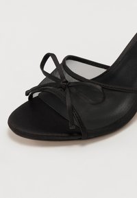 Zwarte high-heeled schoen met een mesh bovenkant, kant detail met strik en een glad satijnen teenstuk. Heeft een slanke, gebogen silhouette.
