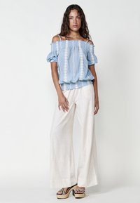 Blusa azzurro chiaro off-the-shoulder con maniche a palloncino e ricami, abbinata a pantaloni bianchi a gamba larga e scarpe con zeppa a righe multicolore.