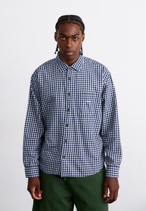 Chemise à carreaux bleu marine et blanche, à manches longues, avec une poche poitrine unique, ourlet rond, boutons bruns, tissu semblant texturé.
