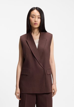 Donna con capelli lunghi e lisci, indossa un blazer senza maniche marrone scuro e pantaloni abbinati su sfondo bianco uniforme.