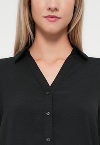 Melna pogu krekls ar V veida apkakli, kam ir gluda tekstūra un sešas priekšpuses pogas. Audumam ir smalka, svītraina uzbūve.