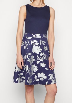 Hihaton laivastonsininen mekko, jossa on vartalonmyötäinen yläosa ja levenevä helma, suuri hopeinen kukkakuosi, yllä seisova nainen.