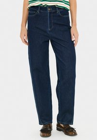 Isik, kes kannab tumeda sinise sirge säärega teksaseid, rohelise ja valge triibuga pluusi ning pruune nahast kingi, seisab valge tausta ees.