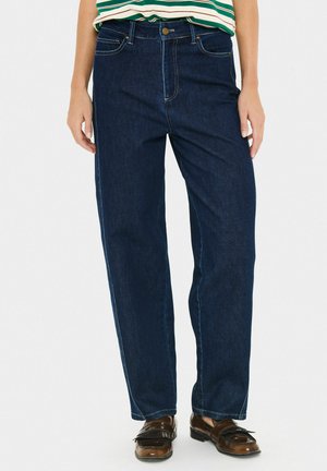 Eine Person trägt eine dunkelblaue gerade geschnittene Jeans, ein grün-weiß gestreiftes Oberteil und braune Lederloafers, steht vor einem weißen Hintergrund.