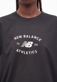 Άτομο που φοράει σκούρο γκρι μπλουζάκι New Balance Athletics με λευκό λογότυπο και κείμενο στο κέντρο του στήθους, με πλεξούδες ορατές στους ώμους.