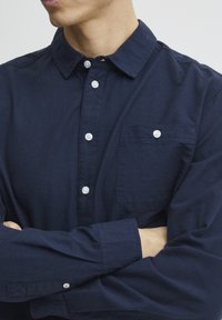 Camicia a maniche lunghe di colore blu navy in cotone, con colletto button-down, tasca frontale e bottoni bianchi, caratterizzata da una texture liscia.