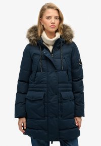 Giacca puffer blu navy con cappuccio foderato in pelliccia, due tasche frontali, cordoncino e dettagli di cucitura verticale. Tessuto morbido, design isolante.