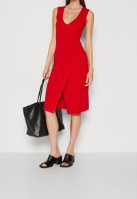 Robe rouge à côtes avec un décolleté en V et un design portefeuille, associée à des mules noires à talons blocs et un sac fourre-tout noir texturé.