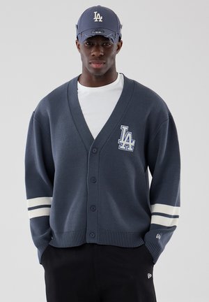 Jeune homme portant une casquette LA bleu marine et un cardigan assorti avec des rayures blanches sur les manches et le logo LA sur la poitrine, t-shirt blanc en dessous.