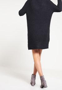 Robe pull en marine foncé, avec une coupe ample et des manches longues. Portée avec des bottines grises, mettant en valeur un tissu texturé.