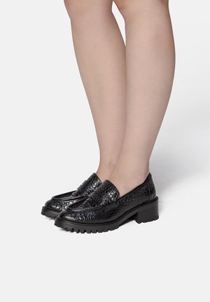Ben iført sorte loafers i krokodilletekstureret læder med chunky lave hæle, stående på hvid baggrund.