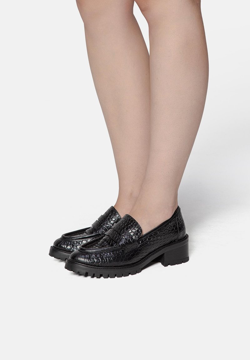 Jambes portant des mocassins en cuir à texture crocodile noire avec des talons bas épais, posées sur un fond blanc.