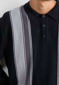 Person mit einem schwarzen Poloshirt, das an der Vorderseite vertikale graue und weiße Streifen hat und drei schwarze Knöpfe nahe dem Kragen.