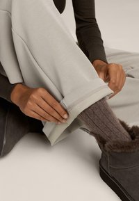 Svetlo sivi hlače s manšetami gladke teksture, prekrivajo rjave pletene nogavice in sivo krzno obložene škornje. Roke prilagajajo manšeto.