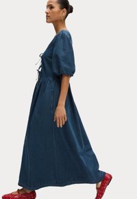 Denim maxi-jurk met pofmouwen, verstelbare taille en zijzakken. Draag met rode klompen met witte stippen.