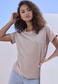 Kurzärmliges, hellbeiges T-Shirt aus weichem Stoff mit Rundhalsausschnitt, getragen über hellblauen Jeans, vor einem klaren blauen Himmel.