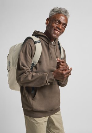 Felpa marrone con cappuccio, zaino beige con dettagli neri e pantaloni chiari. Tessuto morbido con vestibilità rilassata e tasca frontale.