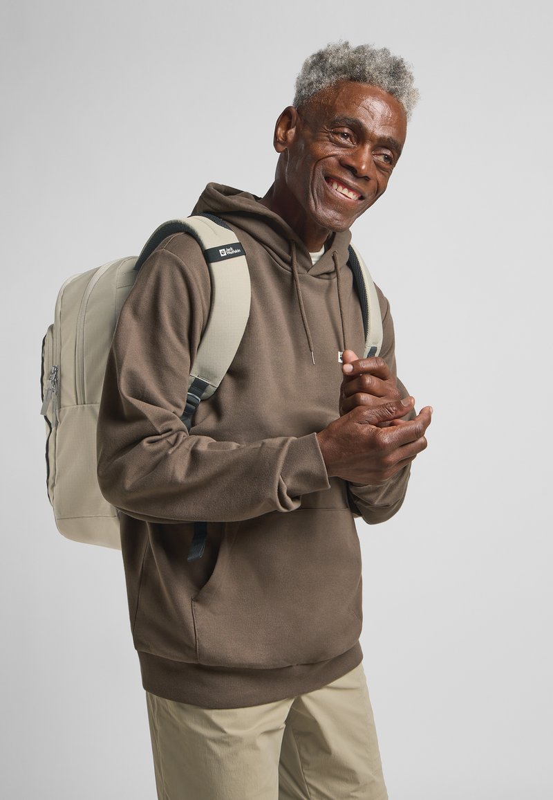 Sudadera marrón con capucha, mochila beige con detalles negros y pantalones de color claro. Tela suave con un ajuste relajado y bolsillo frontal.