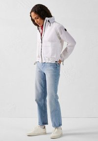 Witte lichtgewicht jas met een rits, twee frontzakken, elastische zoom en verstelbare trekkoorden. Draagt bij lichte blauwe jeans en sneakers.
