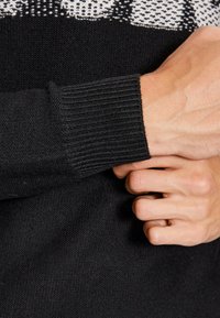 Maglione nero a maglia con polsini a coste, che presenta un motivo testurizzato nella parte superiore con elementi bianchi in contrasto.