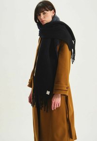 Écharpe noire à franges en matériau doux, portée sur un manteau marron. L'écharpe possède une étiquette en patch près des franges.