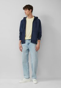 Granatowy sweatshirt z kapturem, jasnożółta koszulka, jasnoniebieskie dżinsy i białe sneakersy. Casualowy strój o luźnym kroju i miękkich teksturach.