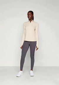 Žena stojaca proti jednoduchému pozadi, oblečená v béžovej dlhé rukávy so zipsom, tmavošedých legínach a bielych teniskách.