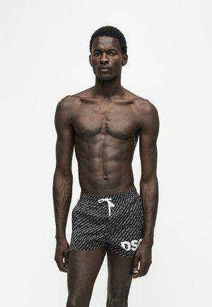 Jeune homme musclé debout, portant un short de bain noir à motifs avec un cordon blanc et des lettres blanches sur la jambe gauche, sur fond blanc uni.