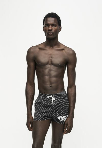 Jeune homme musclé debout, portant un short de bain noir à motifs avec un cordon blanc et des lettres blanches sur la jambe gauche, sur fond blanc uni.