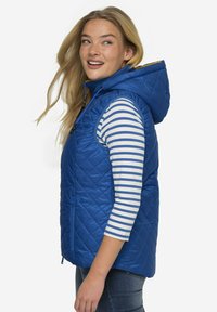 Modrý prošívaný vest s kapucí a předním zipem, zdobený diamantovým vzorem a bočními kapsami. Nositel má pod vestou pruhovanou košili s dlouhým rukávem.