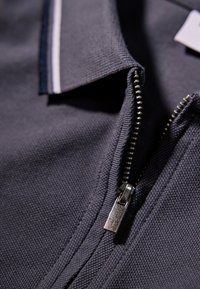 Dunkelgraues Poloshirt aus strukturiertem Stoff mit einem Reißverschluss vorne und weißer Akzentnaht entlang des Kragens und der Knopfleiste.