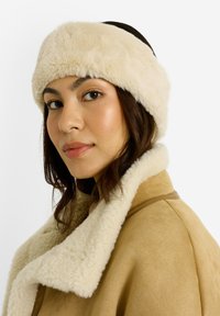 Bandeau en fausse fourrure de couleur crème, avec une texture douce. Le mannequin porte un manteau beige avec un col en fourrure, mettant en valeur une palette chaude.