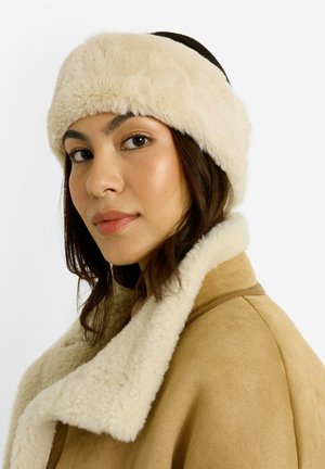 Bandeau en fausse fourrure de couleur crème, avec une texture douce. Le mannequin porte un manteau beige avec un col en fourrure, mettant en valeur une palette chaude.