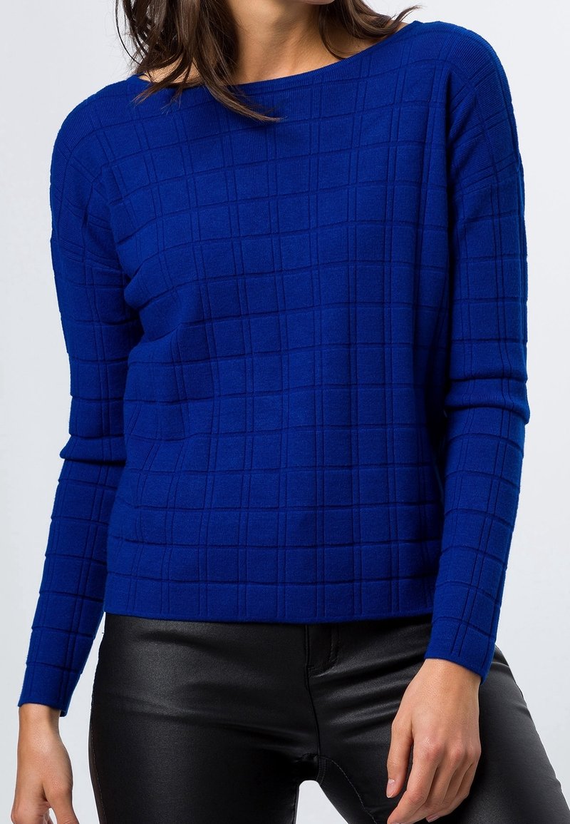 Femme portant un pull texturé bleu roi à manches longues et un pantalon en cuir noir, debout sur un fond clair uni.