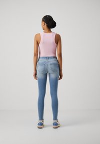 Mujer con moño trenzado que lleva una blusa sin mangas rosa, jeans ajustados de mezclilla azul claro deslavado y zapatillas blancas, de pie de espaldas sobre un fondo liso.