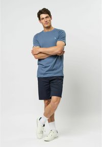 Modré tričko s krátkým rukávem s malou emblematickou nášivkou, doplněné tmavými šortkami. Model v bílých teniskách a ponožkách po kotníky, stojící se založenýma rukama.