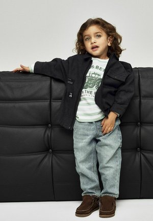 Black zip-up jacket, green striped shirt with tractor graphic, light blue jeans, and brown suede shoes. Child is leaning against a black couch.