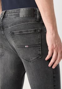 Detailní záběr osoby v černých, vybledlých džínech Tommy Jeans, s viditelnou zadní kapsou a značkovou koženou nášivkou na pase.