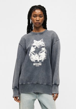 Hosszú fonott hajjal rendelkező nő, aki egy túlméretezett, kopott szürke pulóvert visel, fehér macska grafikával és "MOSCHINO JEANS" felirattal, világos farmerral párosítva.