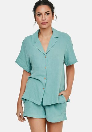Femme portant une chemise à manches courtes bleu clair à boutons et un short assorti en tissu texturé, une main dans la poche, fond neutre.