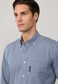 Camisa de botones con un patrón de cuadros en azul y blanco. Presenta un collar puntiagudo y un bolsillo en el pecho con una etiqueta en azul marino. Tela de algodón.