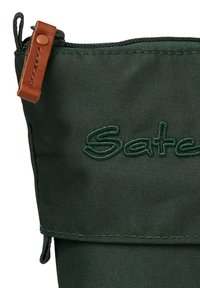 Dunkelgrüner Stoffbeutel mit geprägtem "Satch"-Logo, oberem Reißverschluss und braunem Lederzipper; strukturierte Oberfläche mit sichtbaren Nähten.