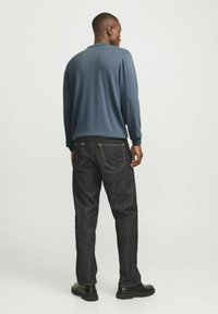 Blaues Langarmhemd, schwarze Jeans mit gelben Nähten und schwarze Schuhe, von hinten vor einem einfarbigen Hintergrund betrachtet.