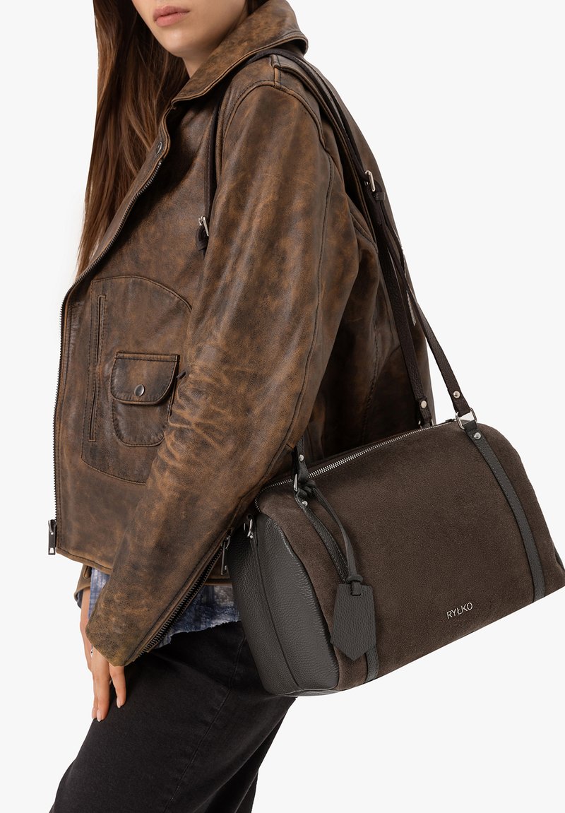 Personne portant une veste en cuir marron tenant un sac à main RYŁKO marron foncé sur l'épaule, devant un fond uni.