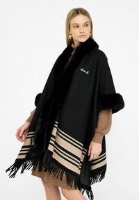 Giovane donna che indossa un poncho frangiato nero e beige con bordo in pelliccia sintetica sopra un abito maglione marrone, mentre guarda di lato.